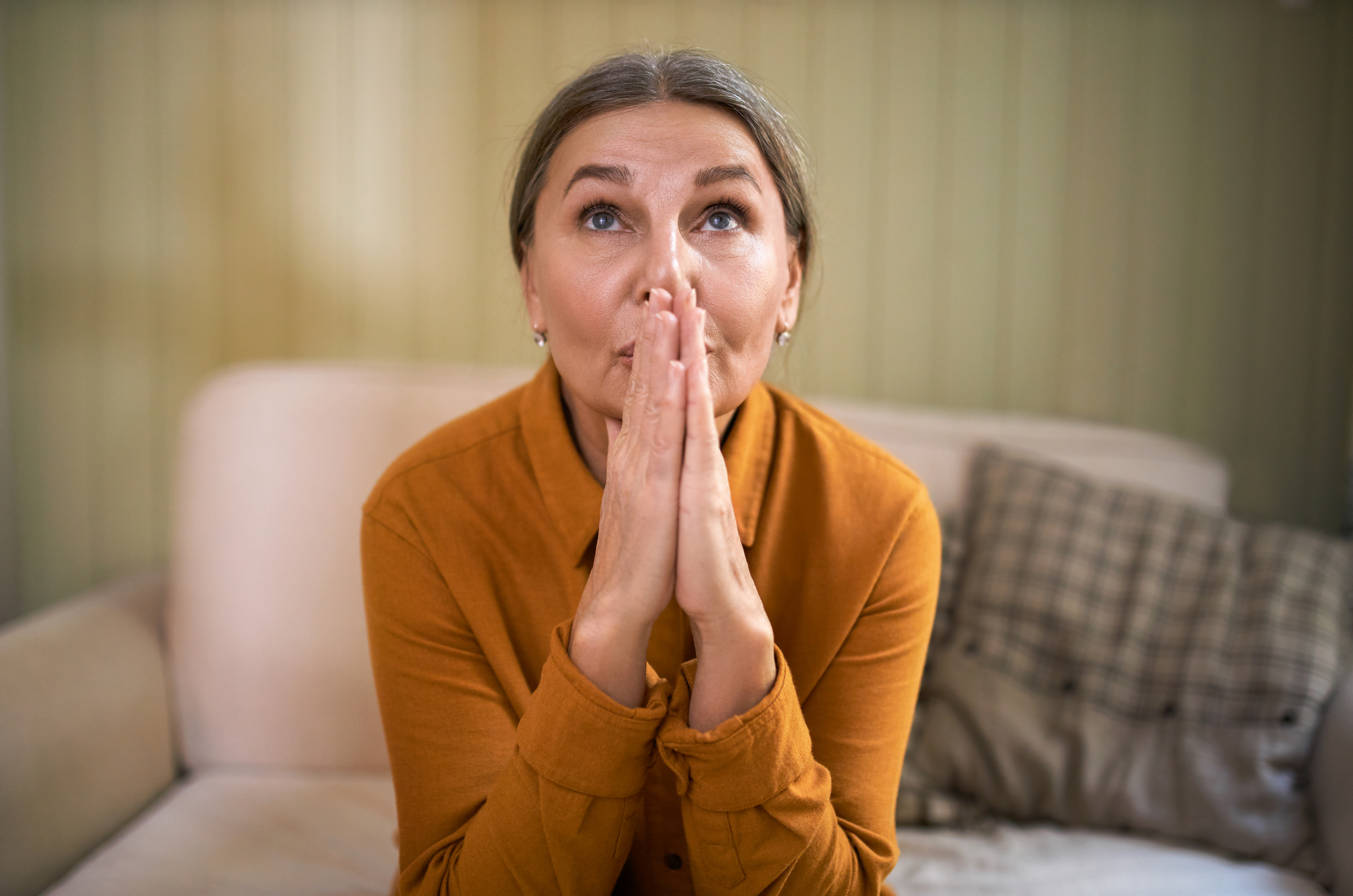 Finding Hope and Strength as a Carent When Feeling Frustrated – A woman in her 50s or 60s wearing a mustard-coloured blouse sits on a sofa, gazing upwards with clasped hands. A moment of resilience, reflection, and finding inner strength in the caregiving journey.